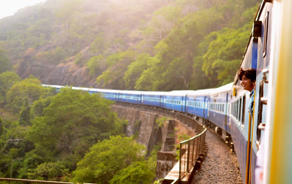 A south-asian girl clings to the handlebars of a train, grinning out the open door at the wild, green forest around her.