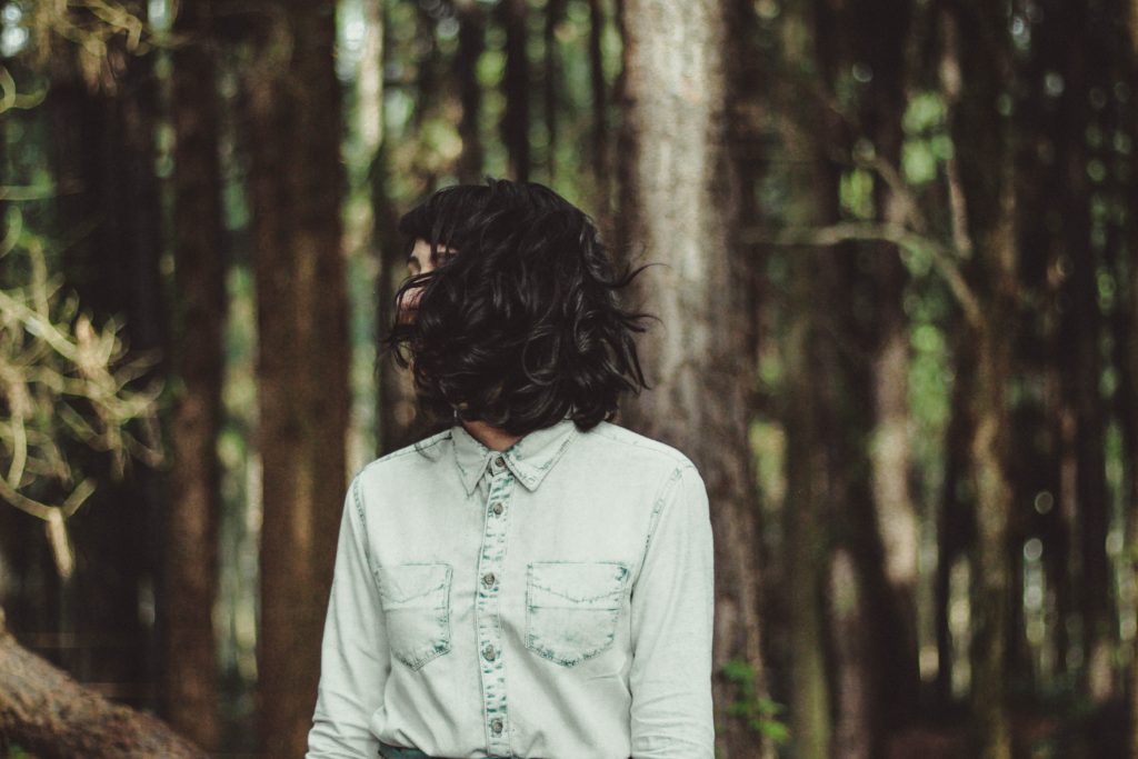 A woman with short hair and a collared, light-wash denim shirt stands in a dark forest.