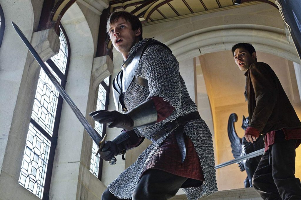 The characters Arthur and Merlin make their way down castle steps, swords in hand, ready to face an enemy