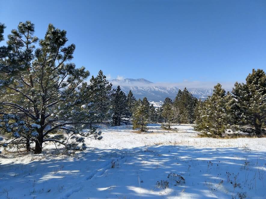 A view of a snow-covered trail lined with evergreens. In the distance, a ribbon of white cloud encircles a range of jagged, snow-speckled mountains. The sky is clear and bright blue.